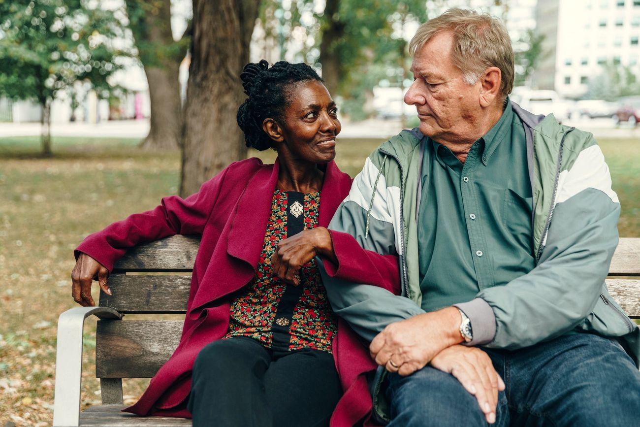 An older couple talking and laughing on a park bench under trees