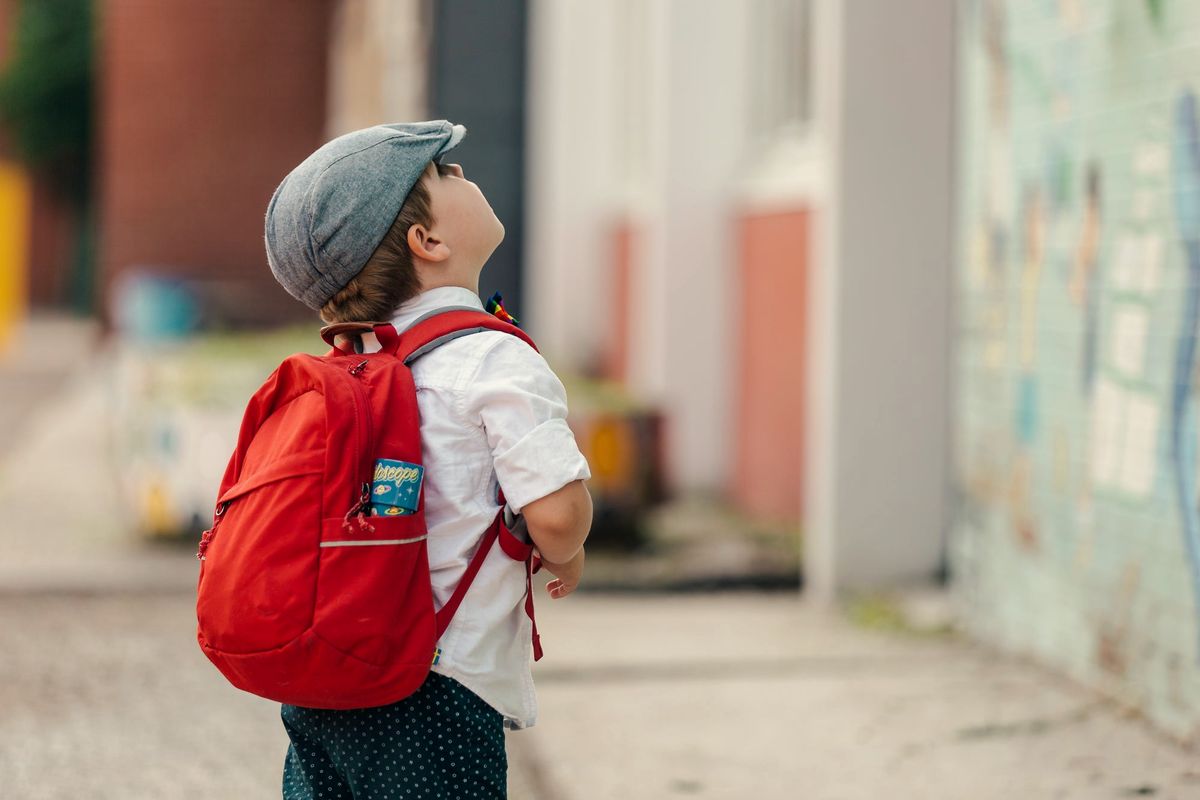 A small boy in a cap and a red backpack looking up at the sky on his way to school