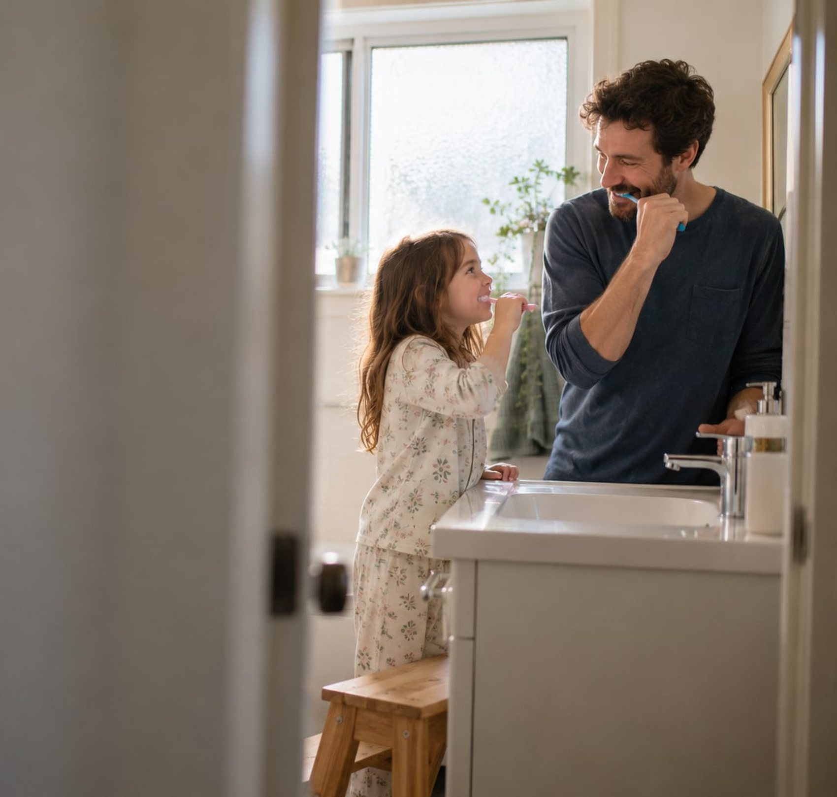 A father and his young daughter brushing their teeth at the bathroom sink in morning light
