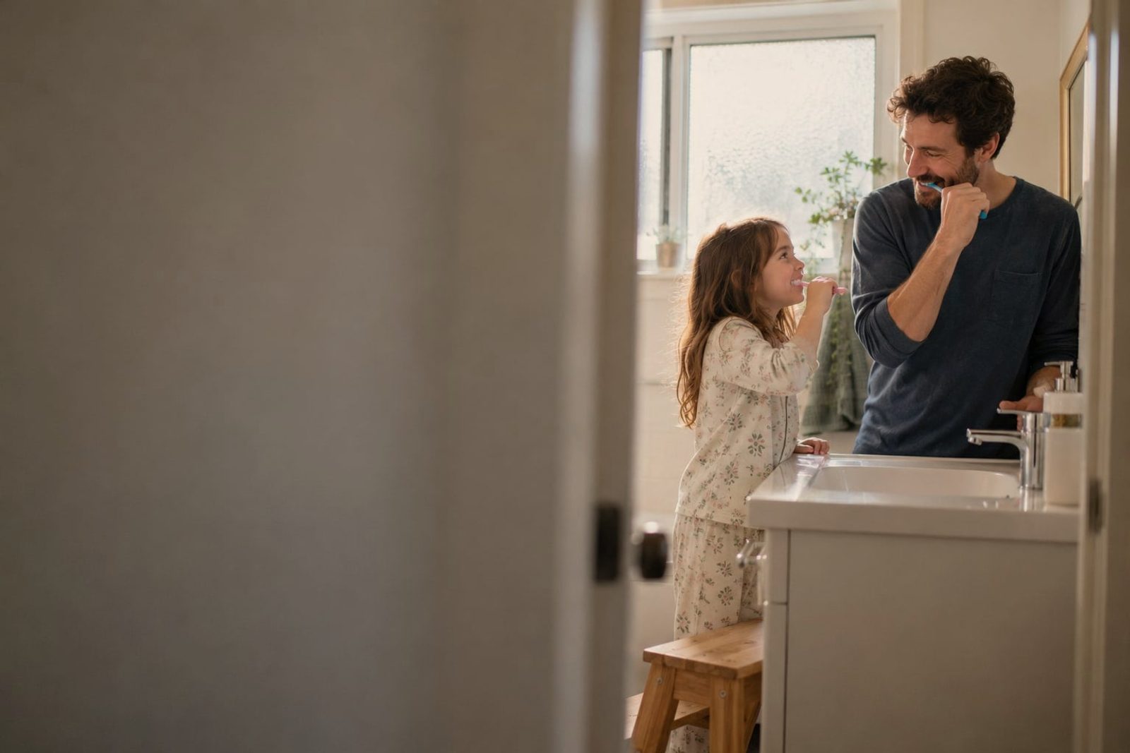 A father and his young daughter brushing their teeth at a bathroom sink in morning light