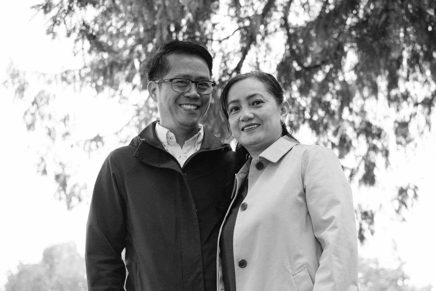 A smiling couple standing close together under trees.