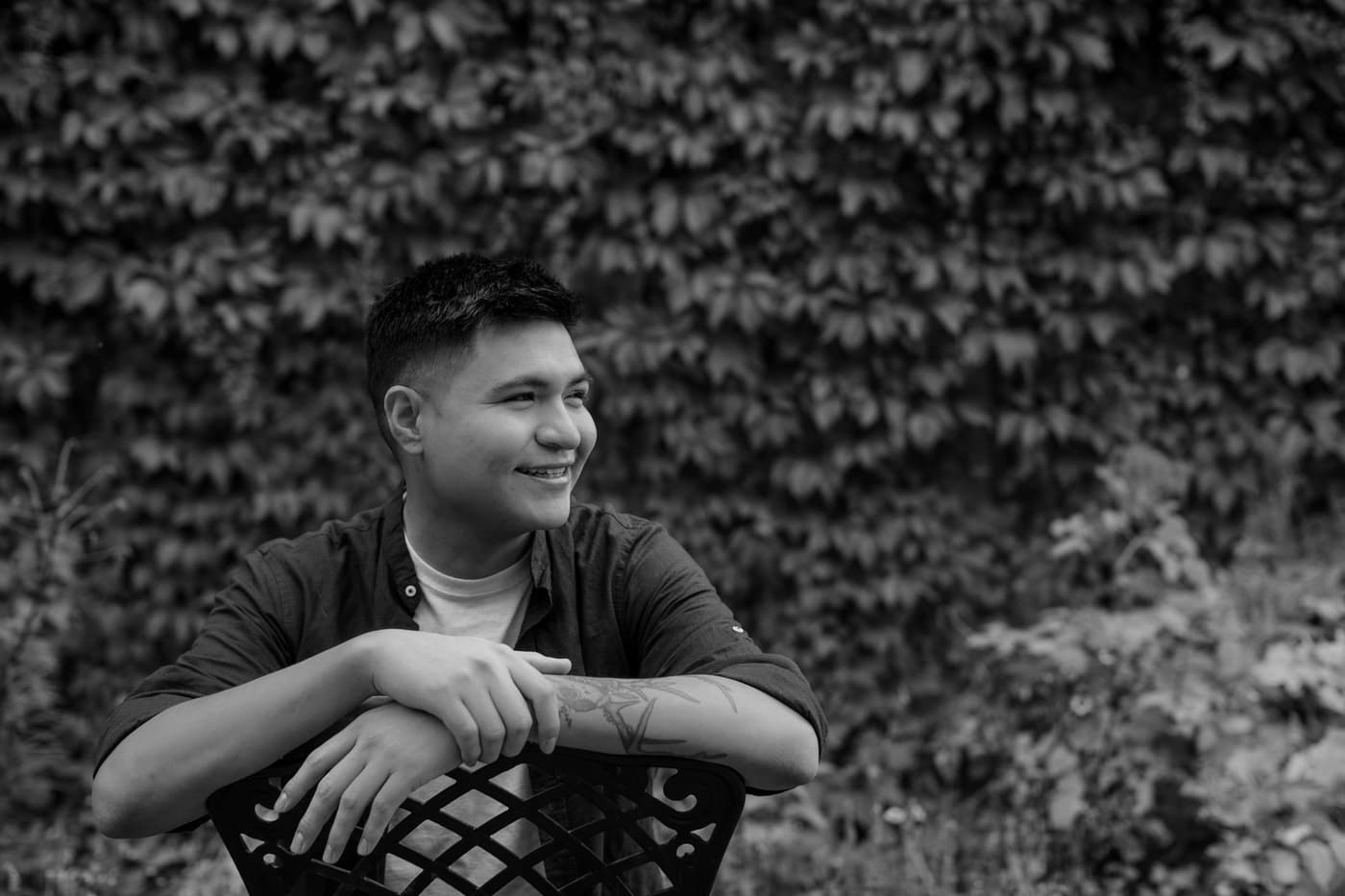 A smiling young man sitting backwards on a chair in front of an ivy wall.