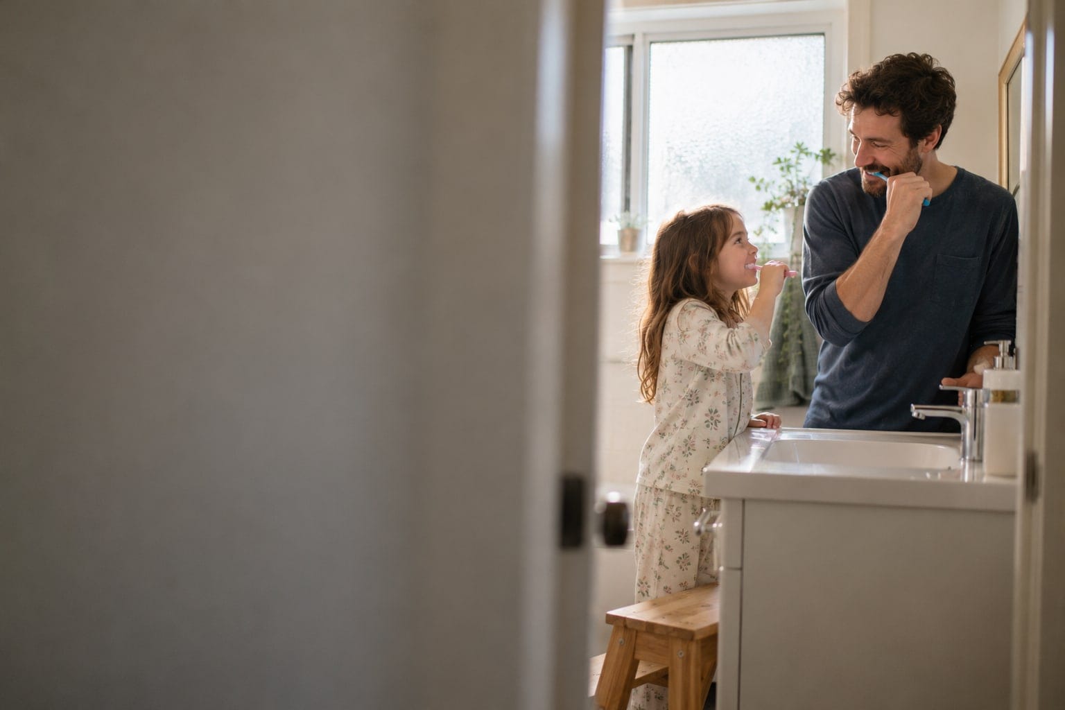 A father and his young daughter brushing their teeth at the bathroom sink