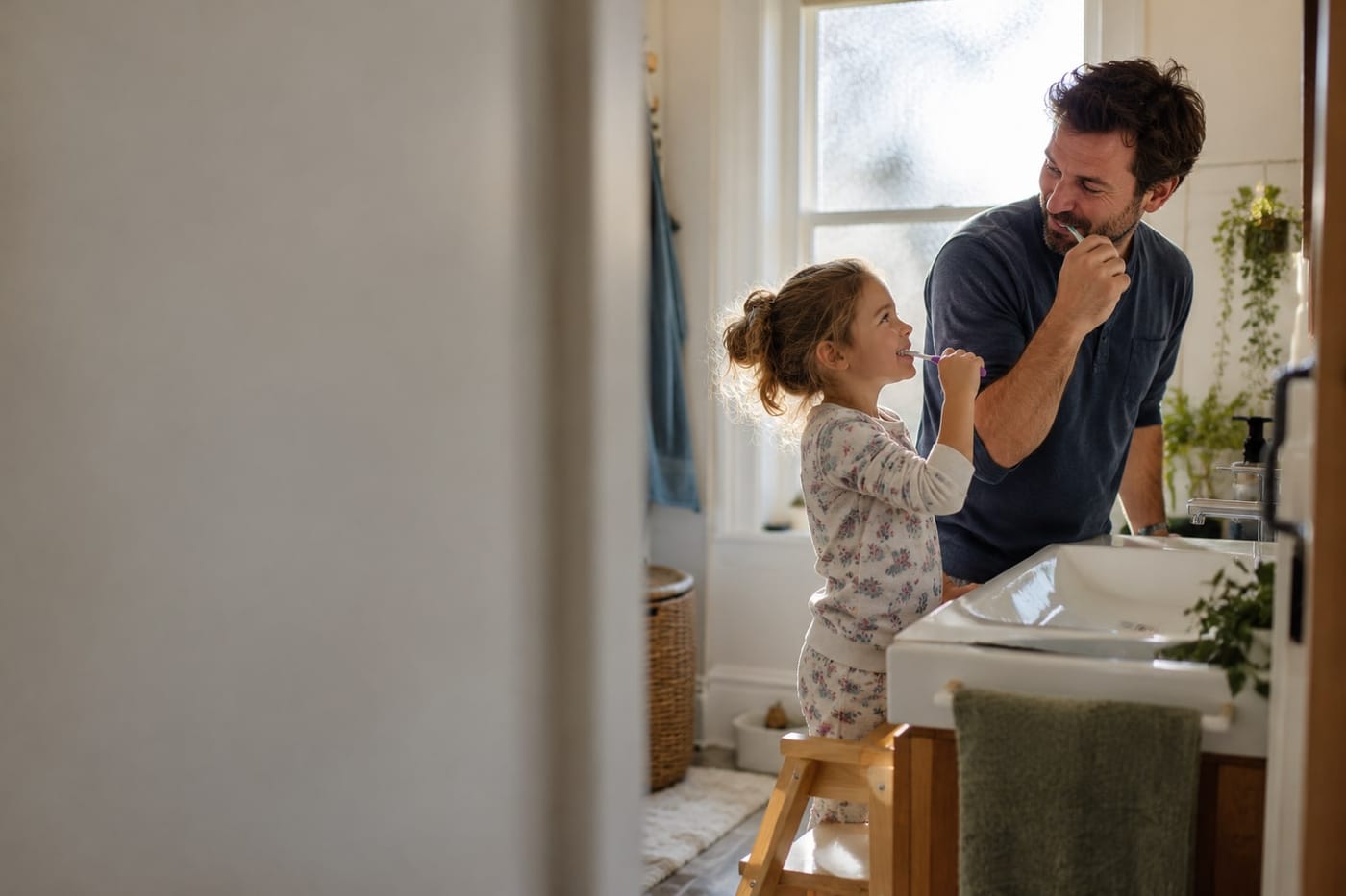 A father and his young daughter brushing their teeth together at the bathroom sink in morning light
